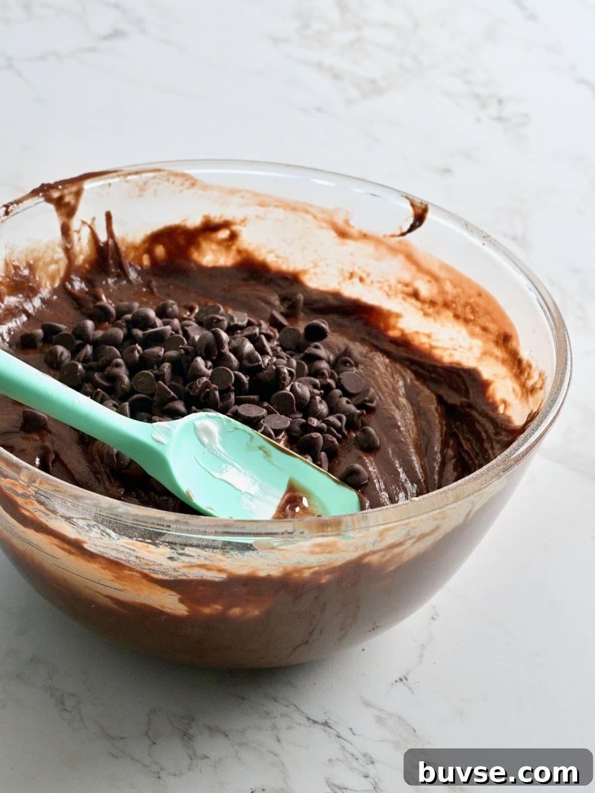 Moist Double Chocolate Chip Bundt Cake 7 Glass bowl of thick chocolate bundt cake batter topped with semi-sweet chocolate chips and a teal spatula.