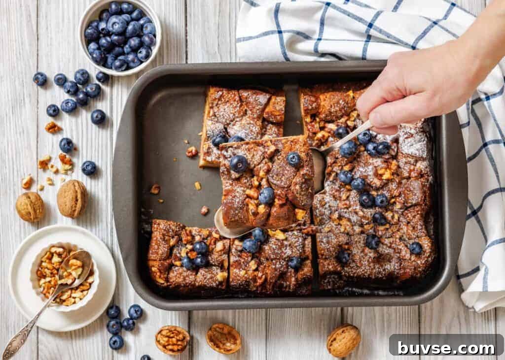A baking pan with sliced Baked Cinnamon French Toast is shown, with a hand using a spoon to lift one piece.