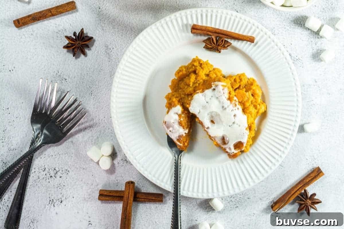 A plate with a serving of sweet potato casserole topped with marshmallows, surrounded by forks, marshmallows, cinnamon sticks, and star anise on a white surface.