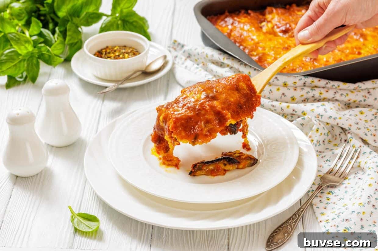 A hand serves a slice of Eggplant Parmigiana onto a white plate, with a casserole dish, herbs, and condiments visible on a white table.