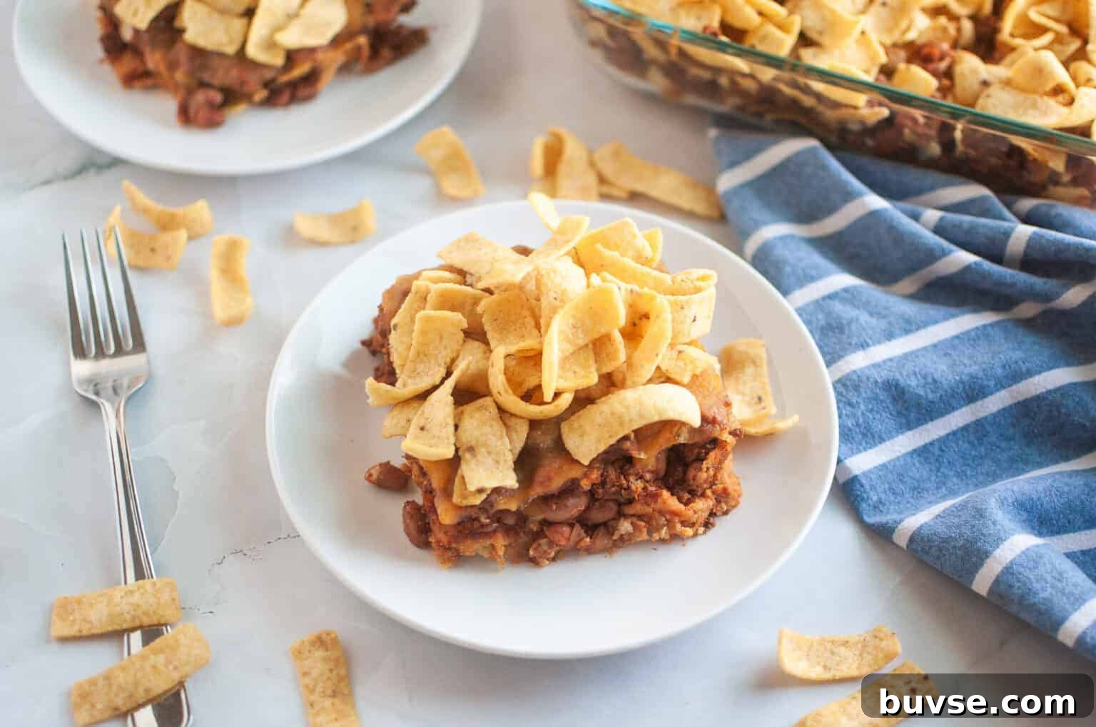 A white plate with a serving of Frito pie, topped with corn chips, is shown on a light surface.
