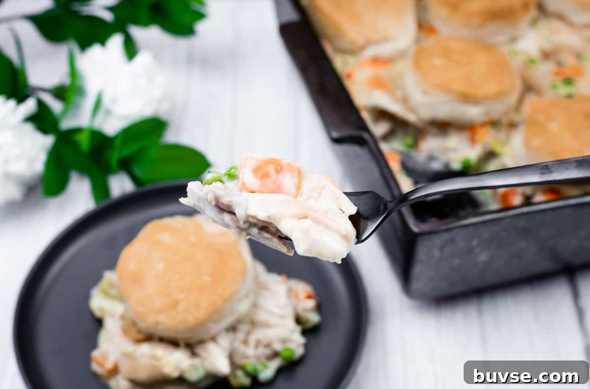 A fork holds a bite of chicken pot pie with vegetables and biscuit topping, with a plate and baking dish of the same meal in the background.