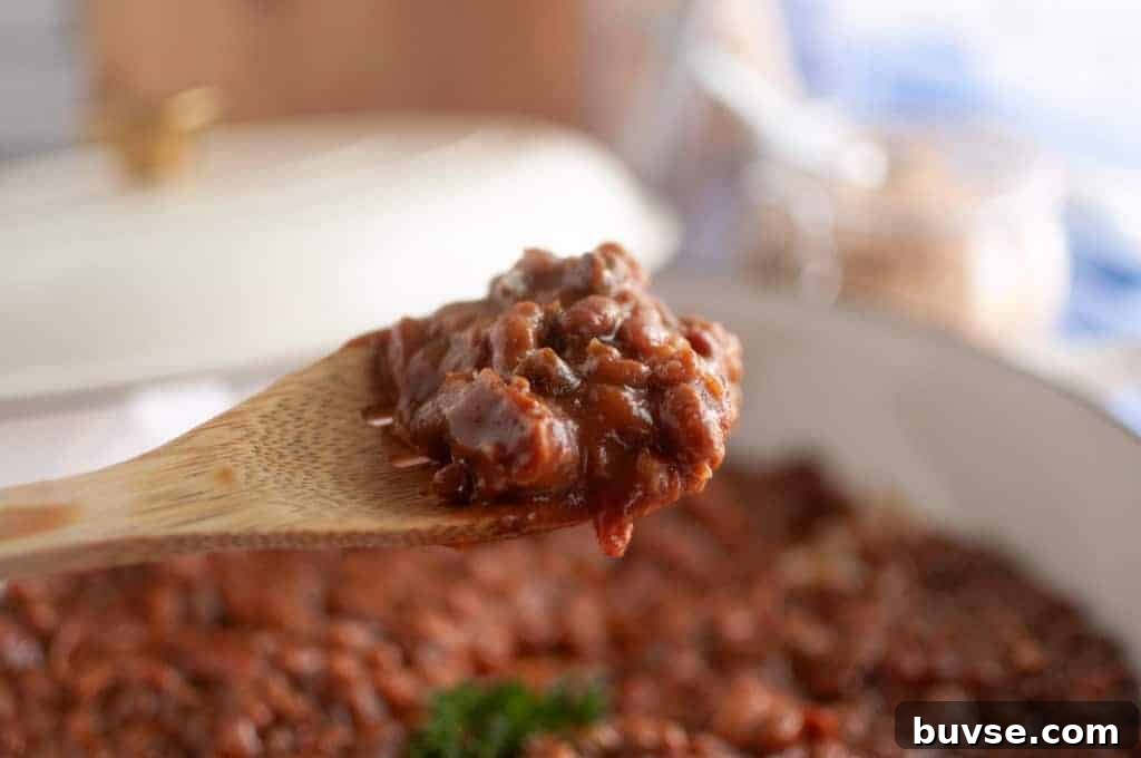 A close-up of a wooden spoon holding a serving of baked beans casserole, with a pot of chili in the background.