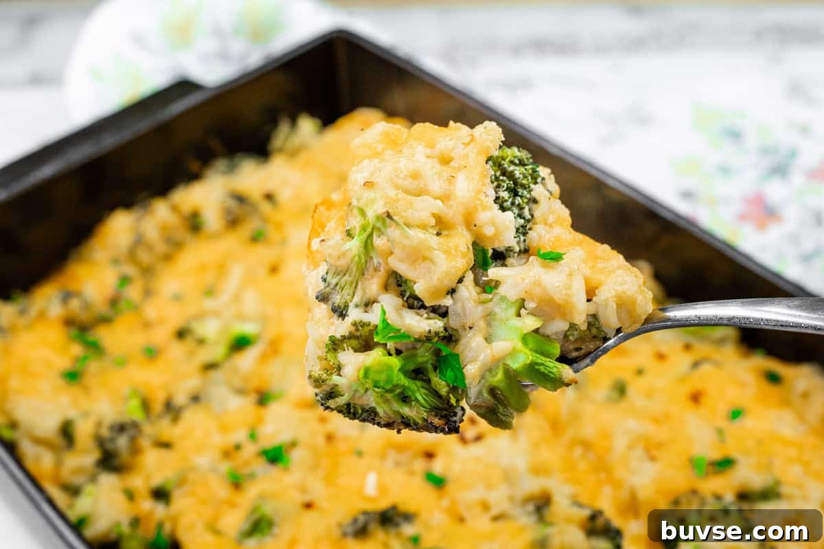 A close-up of a spoon holding a portion of Broccoli Rice Casserole with No Canned Soup, topped with melted cheese, over a baking dish filled with the same dish.