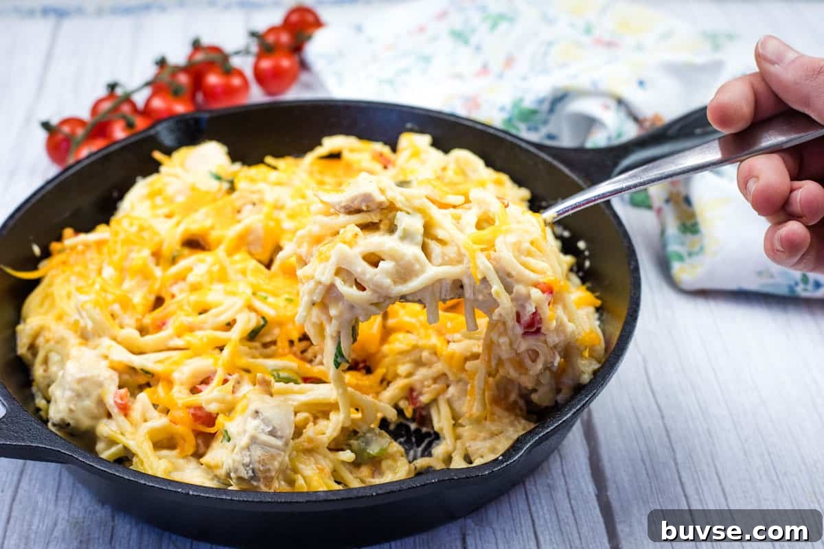 A hand holding a fork with a serving of chicken spaghetti from a cast-iron skillet.