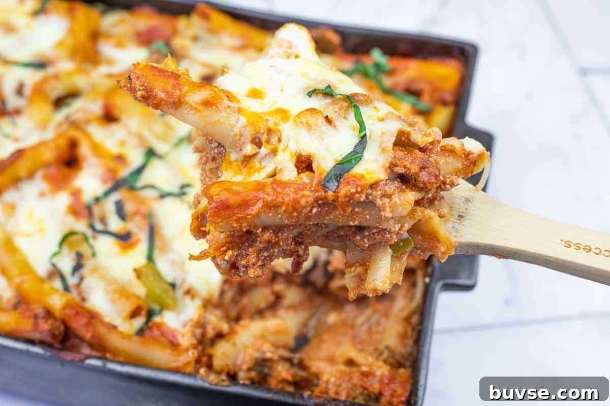 A close-up of a wooden spoon lifting a serving of baked ziti with melted cheese and tomato sauce from a casserole dish.