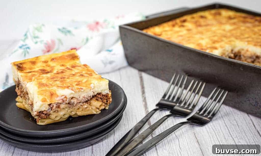 A square piece of pastitsio served on a stack of black plates with three forks and a rectangular baking tray filled with the same pasta on the side.
