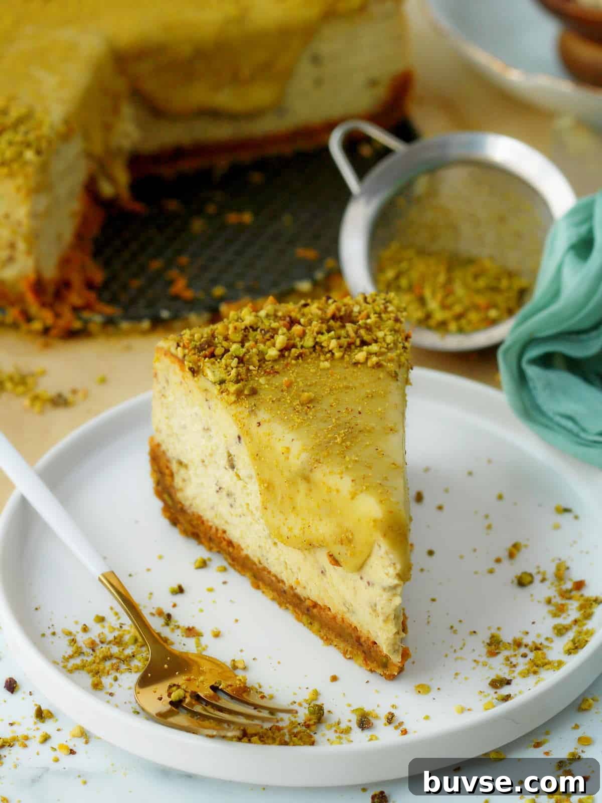 Slice of pistachio cheesecake on white plate, with white fork and whole cheesecake behind it.