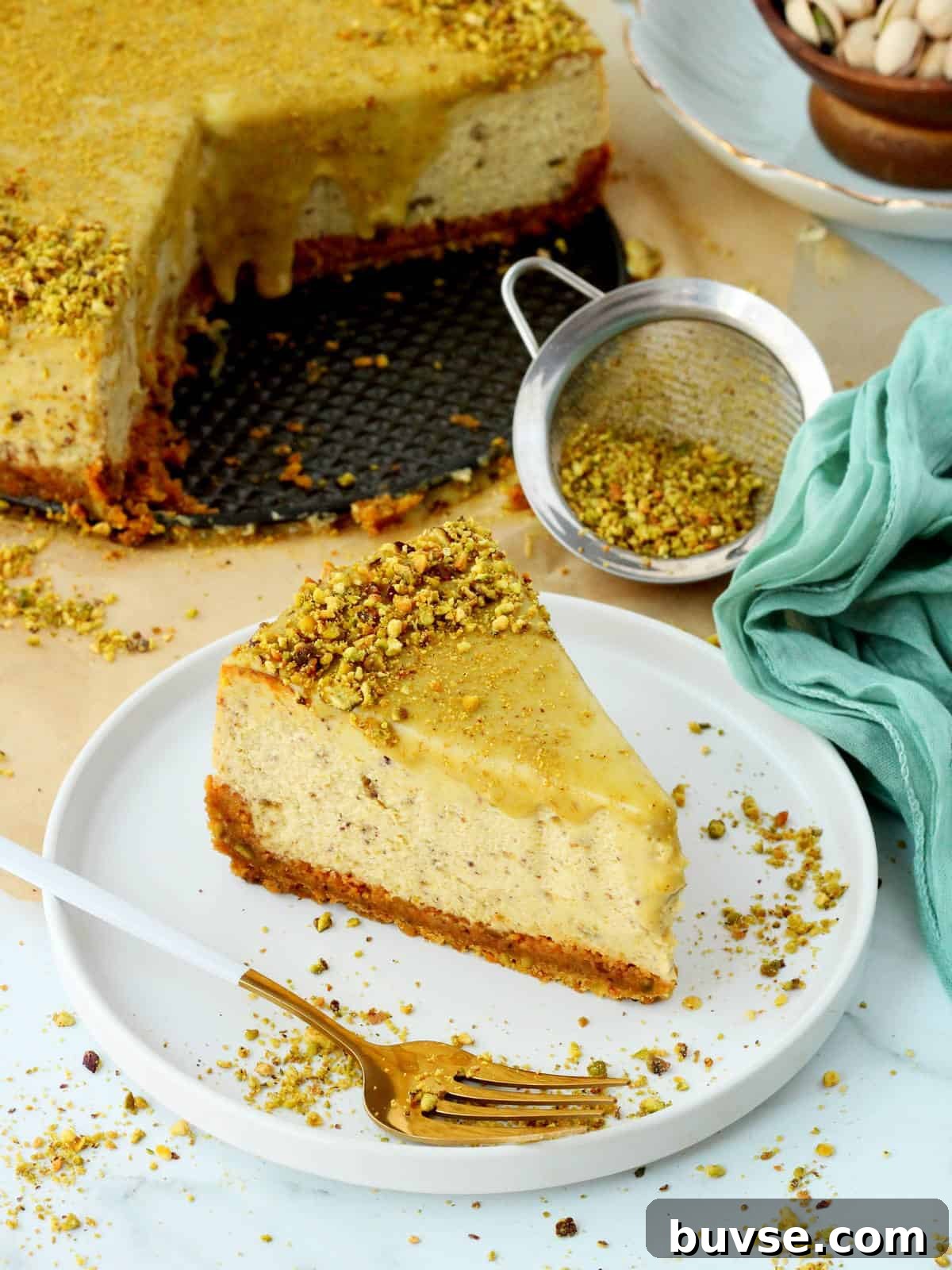 Slice of pistachio cheesecake on white plate, with whole pistachio cheesecake behind it. Green napkin on the right side.