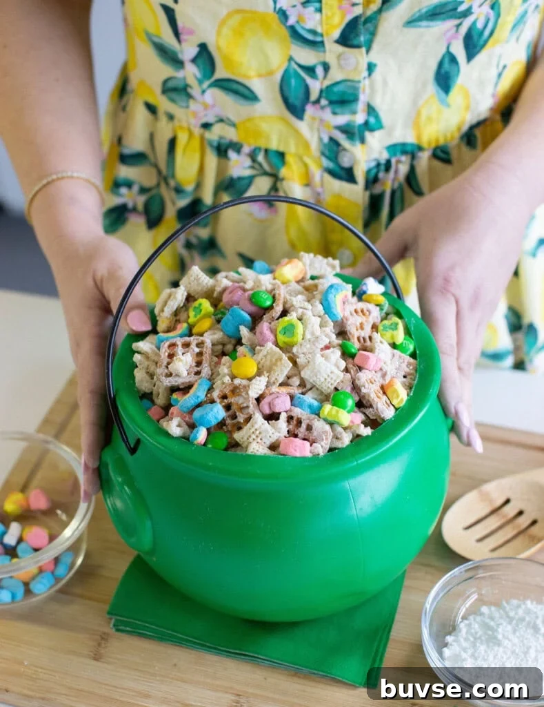 A person in a yellow, lemon-patterned dress holds a green plastic cauldron filled with Chex cereal and colorful marshmallows, with a small bowl of marshmallows and a bowl of powdered sugar nearby.