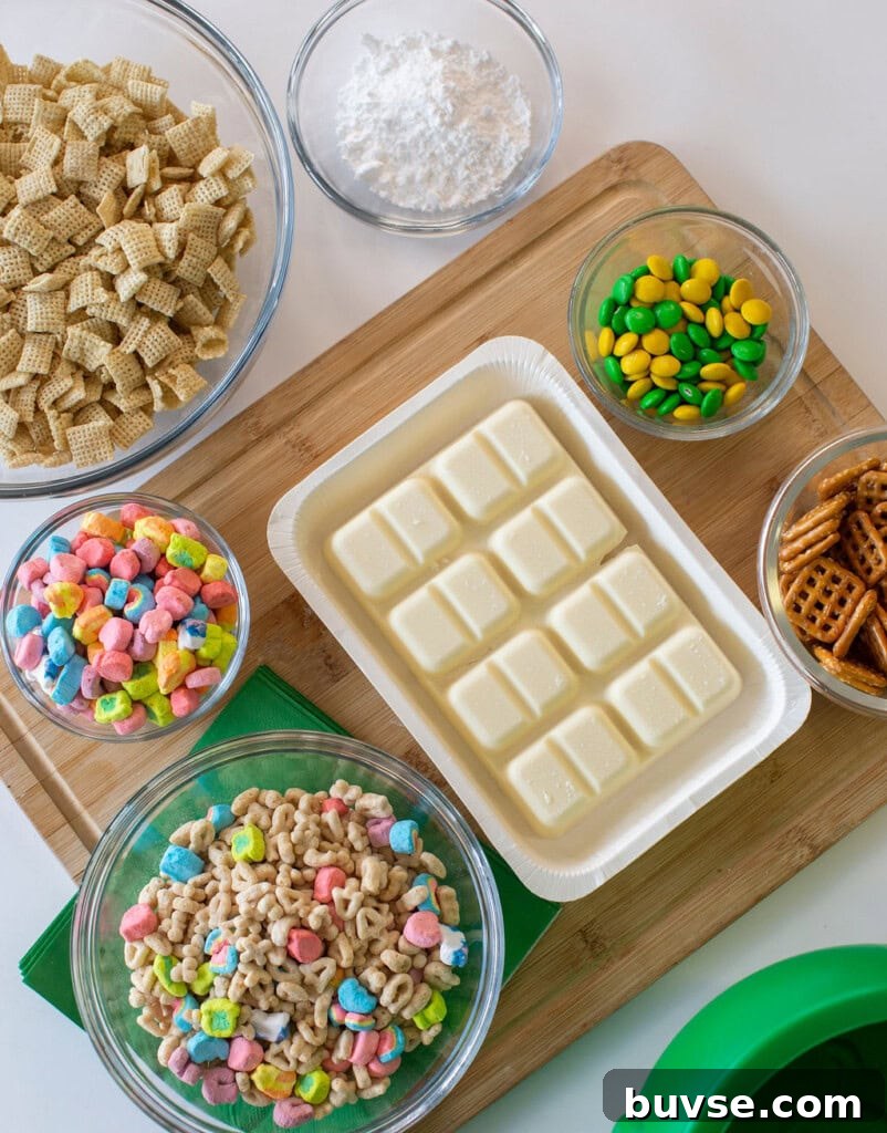 Bowls of Chex cereal, Lucky Charms, pretzels, powdered sugar, and green and yellow candy with a tray of white chocolate on a wooden board, ready for a snack mix recipe.