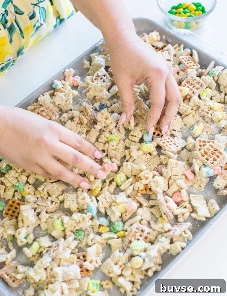 A person arranges a cereal snack mix with colorful candies, pretzels, and assorted cereals on a baking sheet. A small bowl of additional candies sits nearby on a white surface.