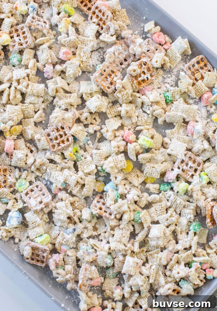 A baking sheet filled with a colorful snack mix of pretzels, Chex cereal, mini marshmallows, coated in a white powdered coating, likely powdered sugar.
