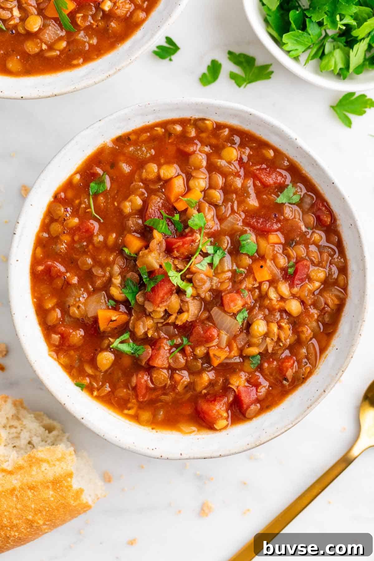 Bowl of vegan lentil soup with a spoon, crusty bread, and fresh parsley on the side.