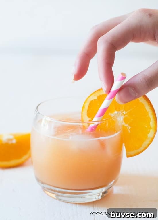 POG Cocktail Recipe: Passion Fruit, Orange and Guava 4 Juice in a glass with an orange garnish. A hand is grabbing the pink and white striped straw.