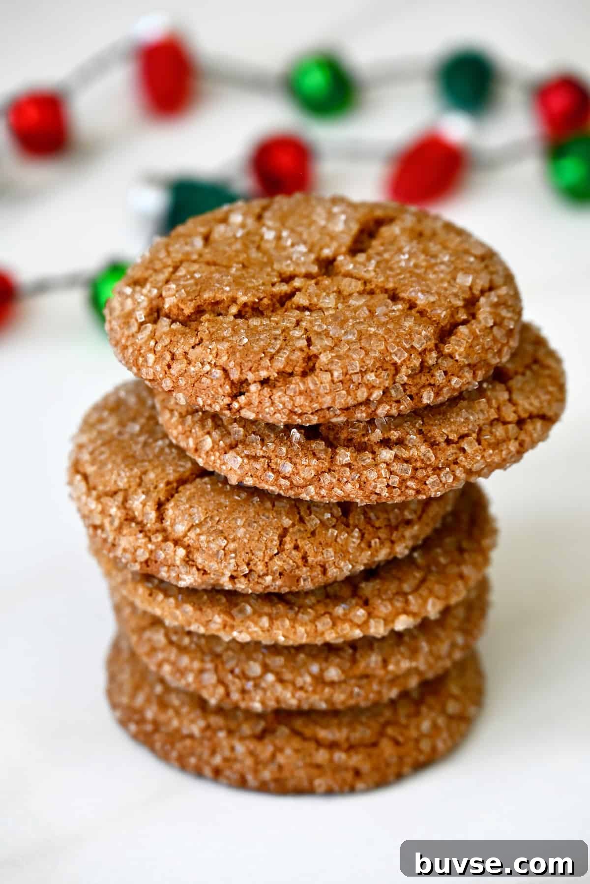Soft Chewy Ginger Cookies Recipe with Molasses and Warm Spices 2 A stack of sugar-dusted ginger cookies are on a white surface, with red and green decorations in the background.
