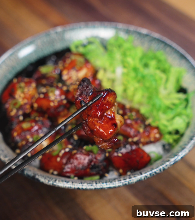 A juicy piece of garlic soy glazed chicken is held up with chopsticks over a bowl filled with caramelized chicken thighs, fluffy rice, and fresh shredded lettuce. The glossy, sticky glaze clings to the chicken, highlighting its crispy edges and tender inside, making the bite look irresistible and packed with flavor.