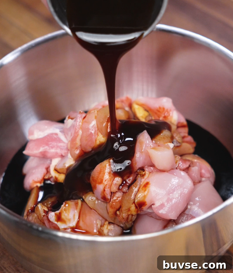 Thick garlic soy marinade is poured from a metal bowl onto bite-sized raw chicken thighs, ready to be marinated. The glossy dark sauce coats the meat, preparing it for a flavorful soak that infuses the chicken with sweet, savory, and garlicky depth before cooking.