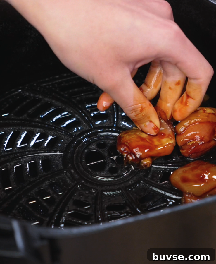 A hand carefully places marinated chicken thighs into an air fryer basket, spacing them apart for even cooking. The chicken is already coated in a glossy garlic soy glaze, showing the first step in achieving the crispy and sticky finish of the final dish.