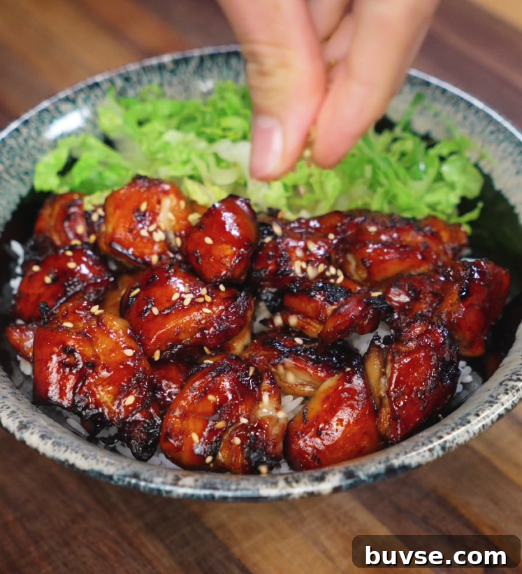 A hand sprinkles sesame seeds over a freshly cooked bowl of garlic soy glazed chicken. The chicken pieces are deeply caramelized with crisp edges and glistening glaze, sitting atop white rice with a side of crisp lettuce, creating the perfect final touch for texture and visual appeal.