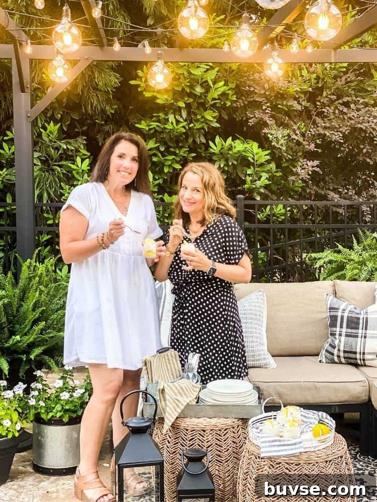 Refreshing Summer Picnic Menu Ideas to Wow Guests 3 Two women holding cups of lemon bars in a jar standing under a pergola strung with different sizes of string lights. The background is all kinds of shrubs. A tan outdoor sofa sits under the pergola with wicker tables, lanterns, and potted plants.