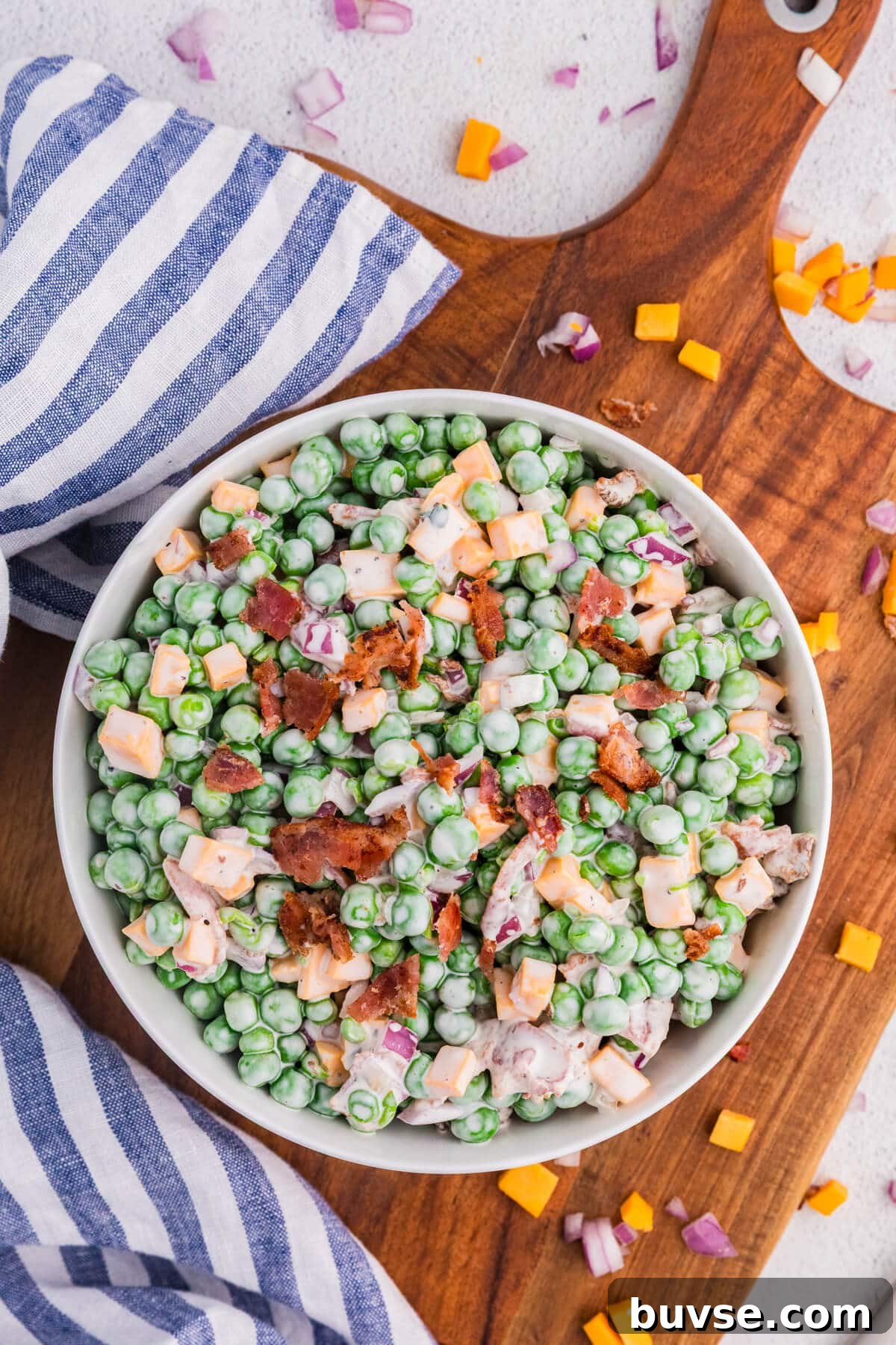 Overhead view of creamy pea salad with green peas, cheddar cheese cubes, crispy bacon, and red onion in a white bowl on a wooden cutting board with striped cloth, fresh summer side dish.