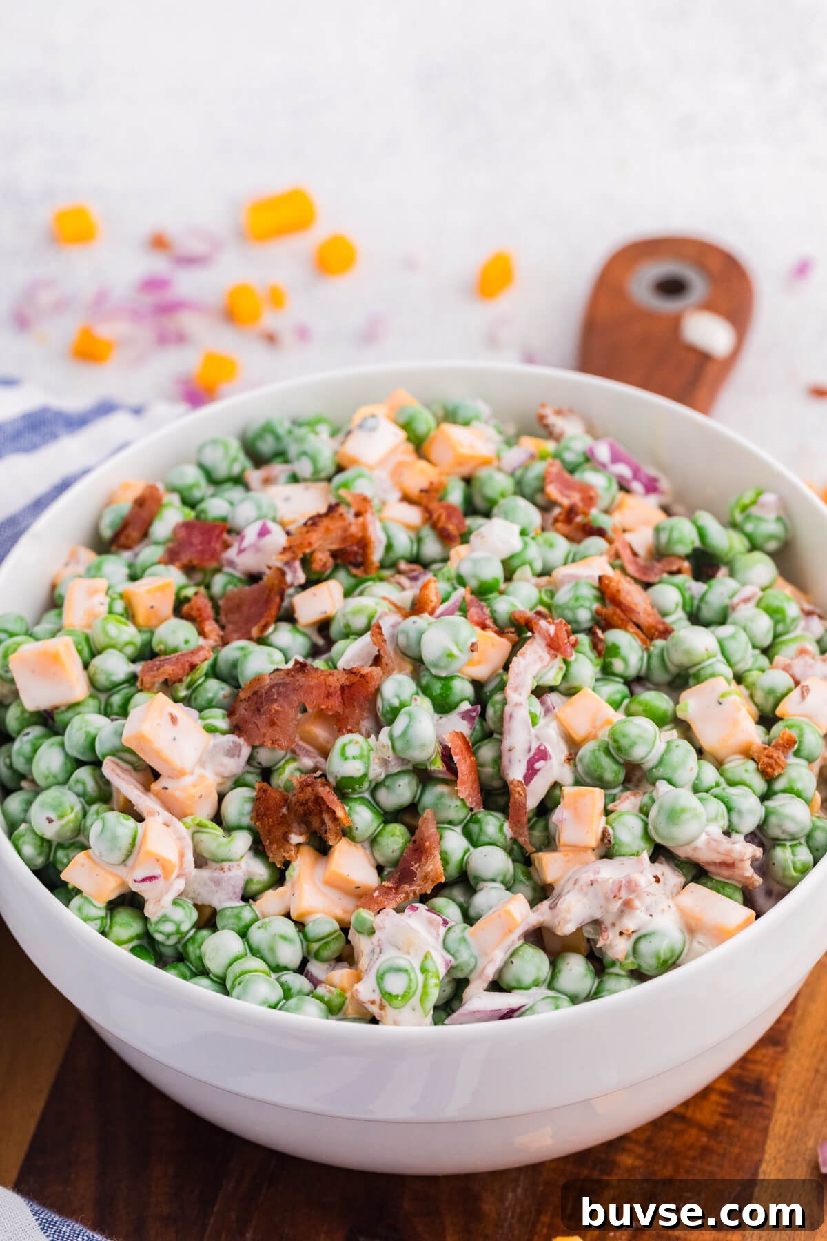 Close-up angled shot of pea salad with creamy dressing, green peas, cheddar cubes, bacon bits, and diced red onions in a white bowl, rustic kitchen background with scattered ingredients.