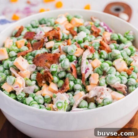 Side view of creamy pea salad in a white bowl featuring green peas, cheese cubes, bacon, and red onion, served on a wooden board with soft blurred background.