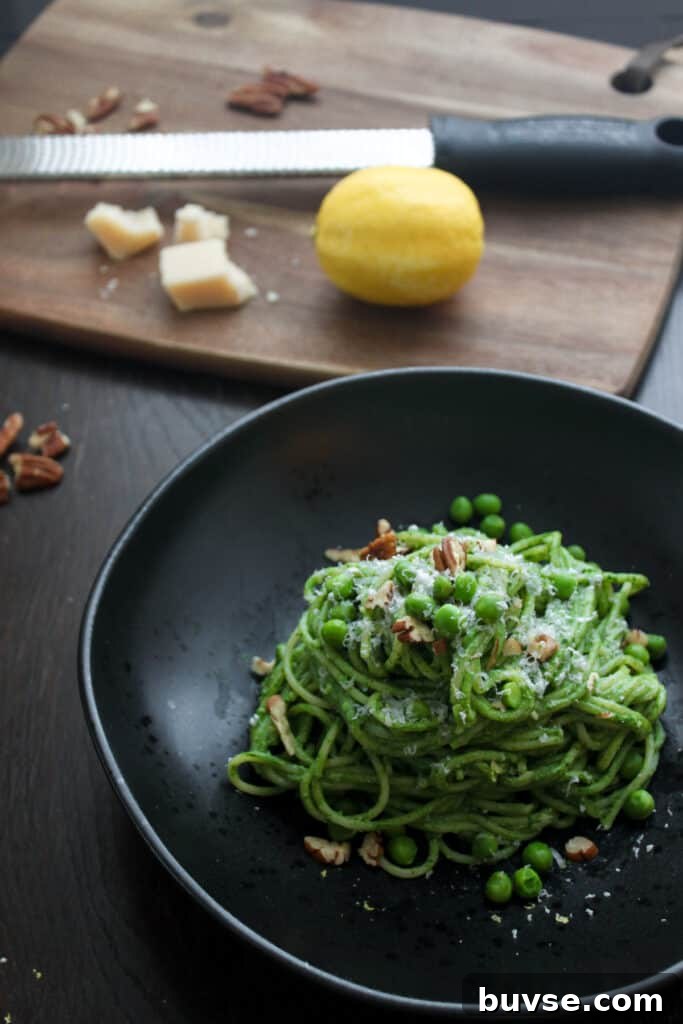 A single portion of creamy spinach pasta on a round black bowl topped with grated parmesan cheese, chopped toasted pecans and green peas. In the background out of focus is a cutting board with a microplane grater, a lemon and parmesan cheese.