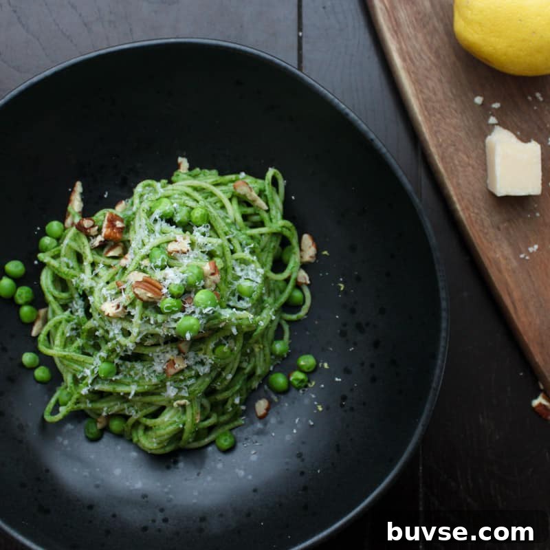 An overhead shot of a single portion of creamy spinach pasta in a black bowl topped with grated parmesan cheese, chopped toasted pecans and green peas. Just to the side is a cutting board with a lemon and parmesan cheese.