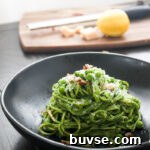 A single portion of creamy spinach pasta on a round black bowl topped with grated parmesan, toasted chopped pecans and green peas. Out of focus is a cutting board with a micrpplane grater, a lemon and parmesan cheese.