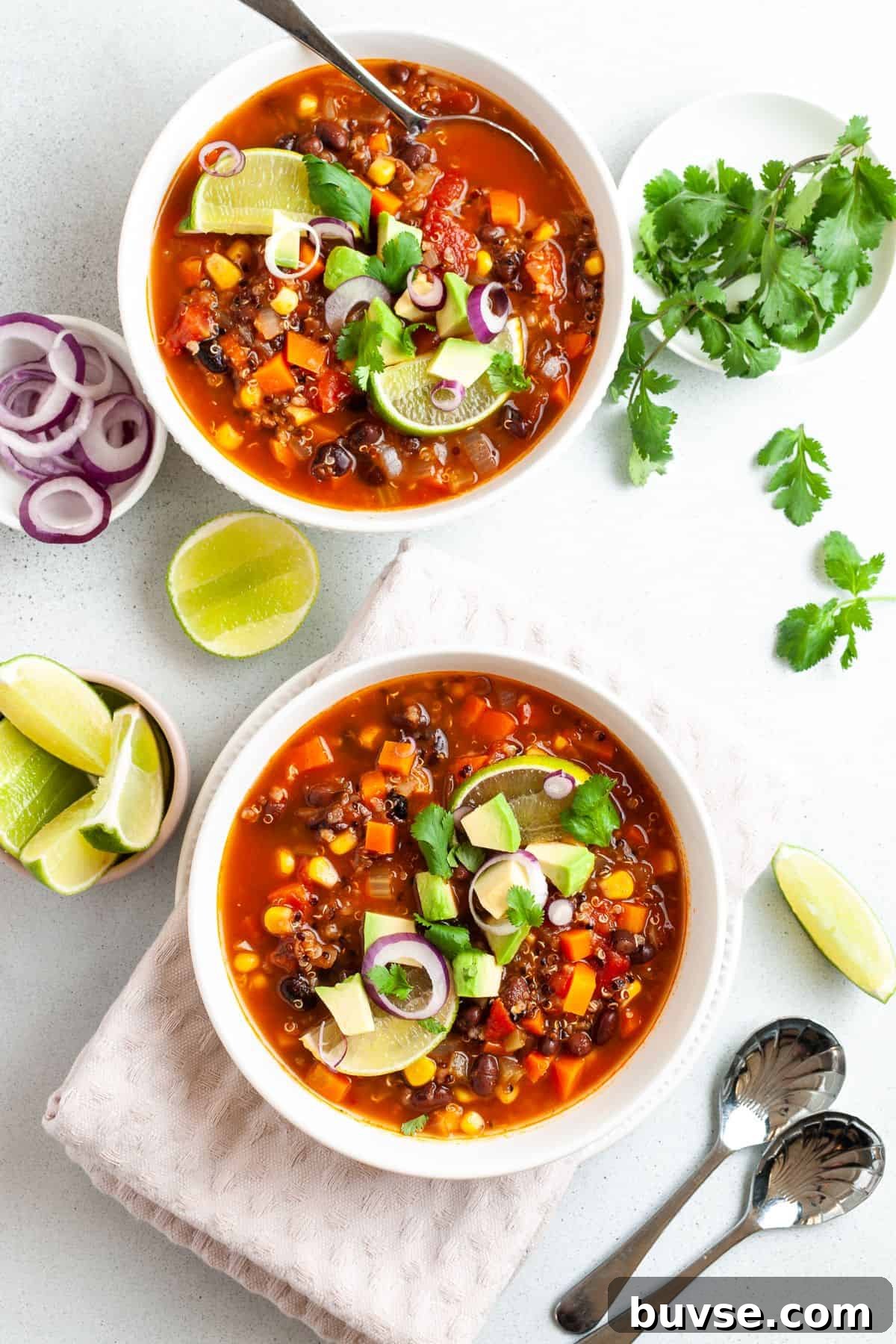 Two round white bowls of soup, one with a spoon in it, with some lime wedges and coriander around the edge of the dishes.