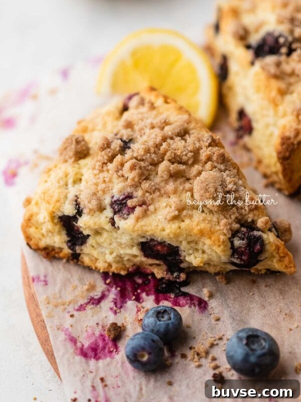 Bakery-Style Lemon Blueberry Muffins with Streusel Topping 15 Lemon blueberry scones with streusel topping placed on a parchment paper lined round wooden serving plate with lemon wedges and blueberries placed around it.