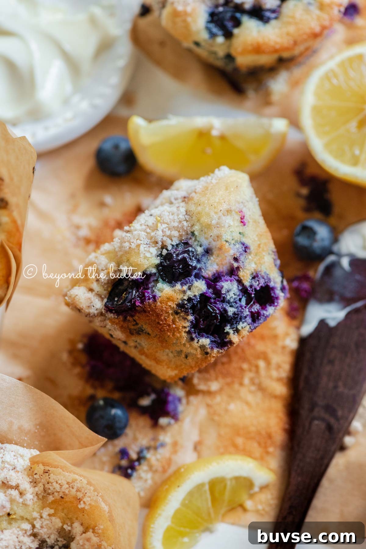 Bakery-Style Lemon Blueberry Muffins with Streusel Topping 4 Unwrapped lemon blueberry streusel muffin with small bowl of butter, lemon wedges and blueberries around it | All images © Beyond the Butter®