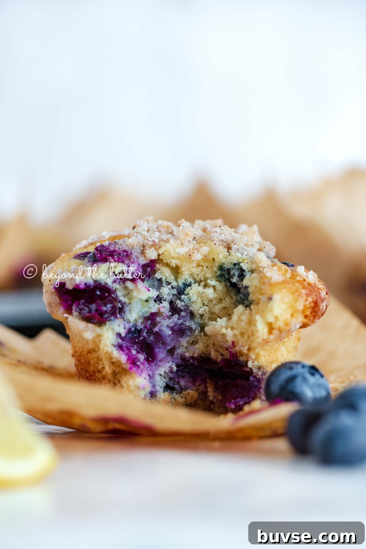 Bakery-Style Lemon Blueberry Muffins with Streusel Topping 9 Unwrapped and half eaten lemon blueberry streusel muffin with lemon wedges and blueberries around it | All images © Beyond the Butter®