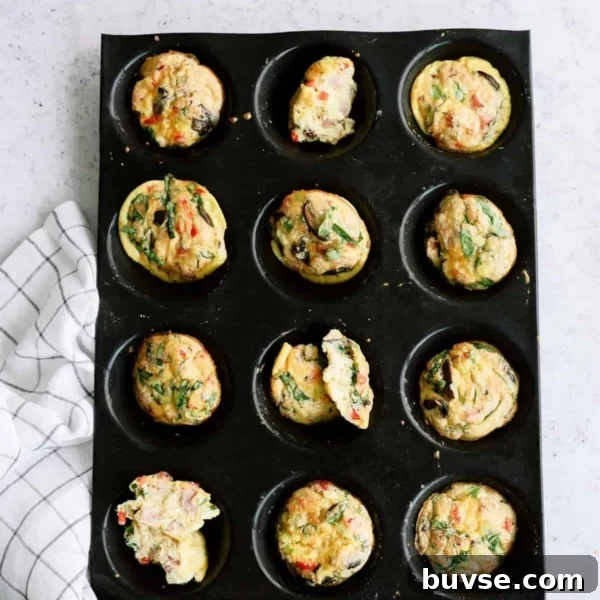 Muffin tray with twelve cooked egg muffins, some with spinach and vegetables, on a light surface next to a checkered cloth.