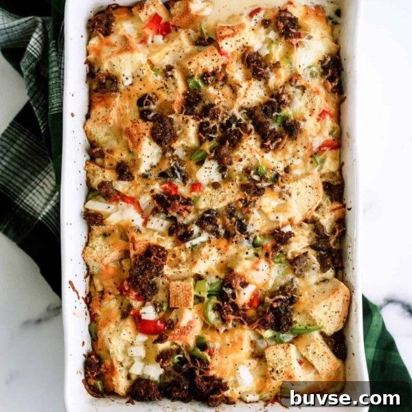 Baked casserole with bread cubes, bell peppers, onions, melted cheese, and browned sausage in a white rectangular dish on a green checkered cloth.