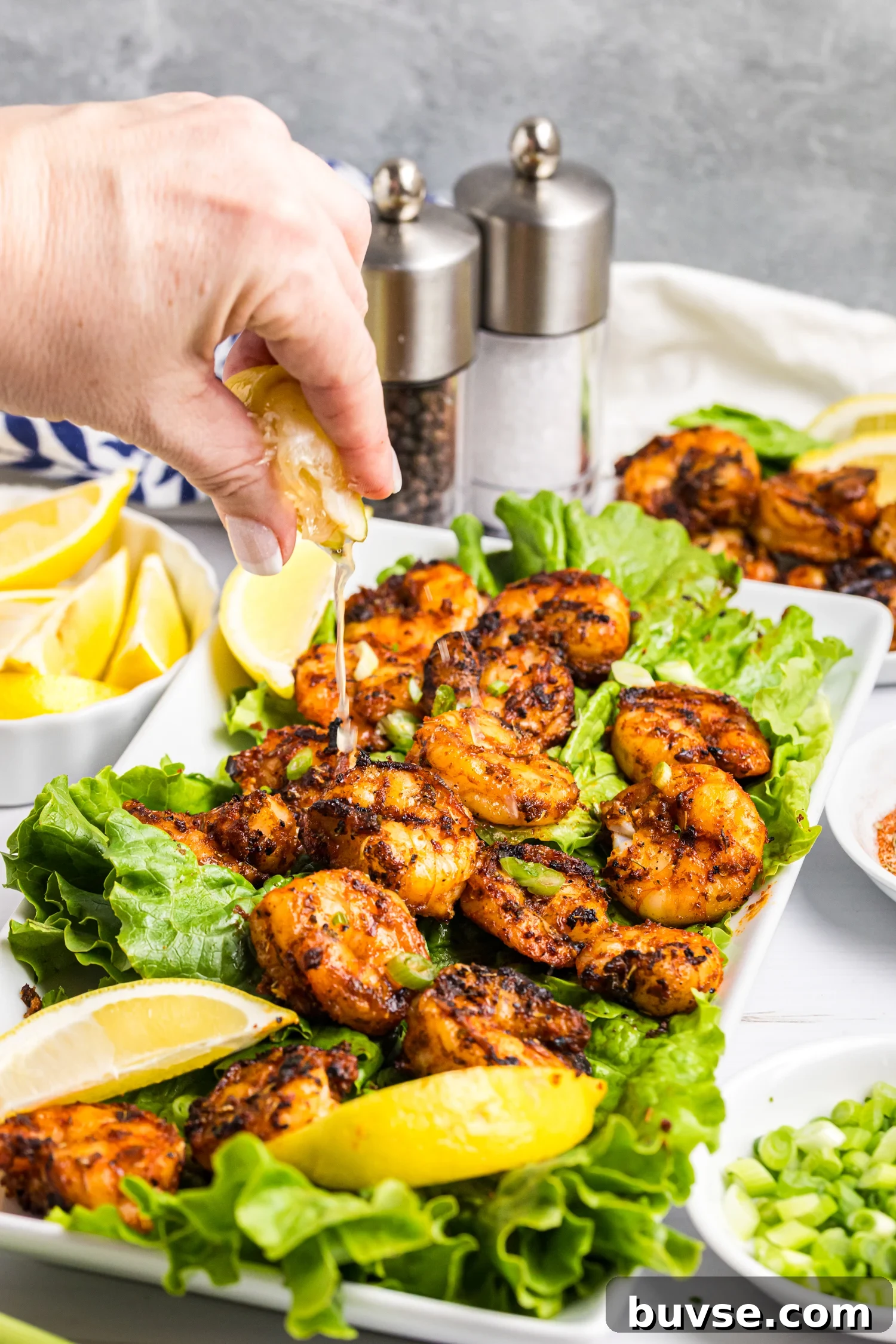 Hand squeezing lemon juice over grilled shrimp served on lettuce with lemon wedges and green onions.