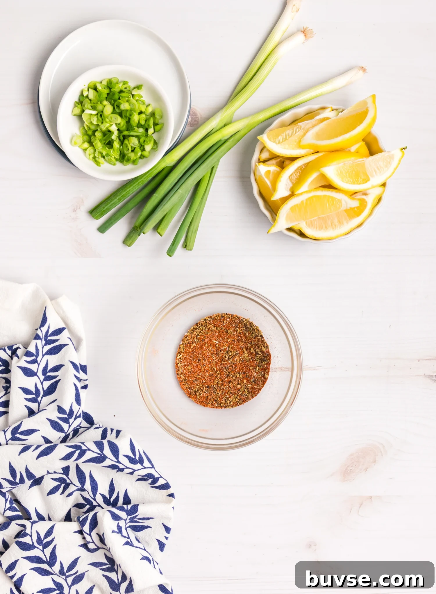 Chopped green onions, whole scallions, lemon wedges, and a bowl of spice blend arranged on a white surface.