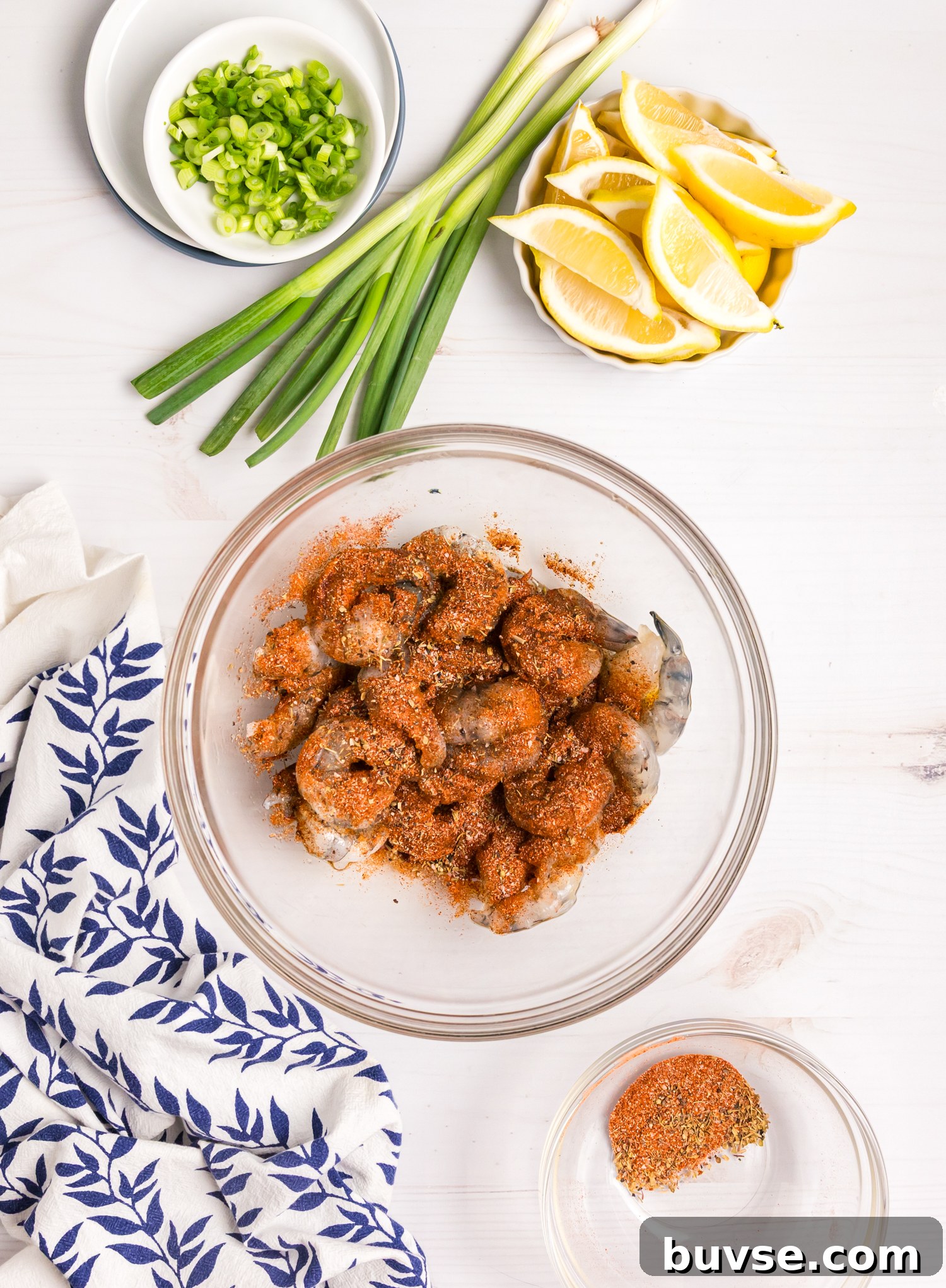 Raw shrimp in a glass bowl coated with a reddish spice mixture, with green onions and lemon wedges nearby.