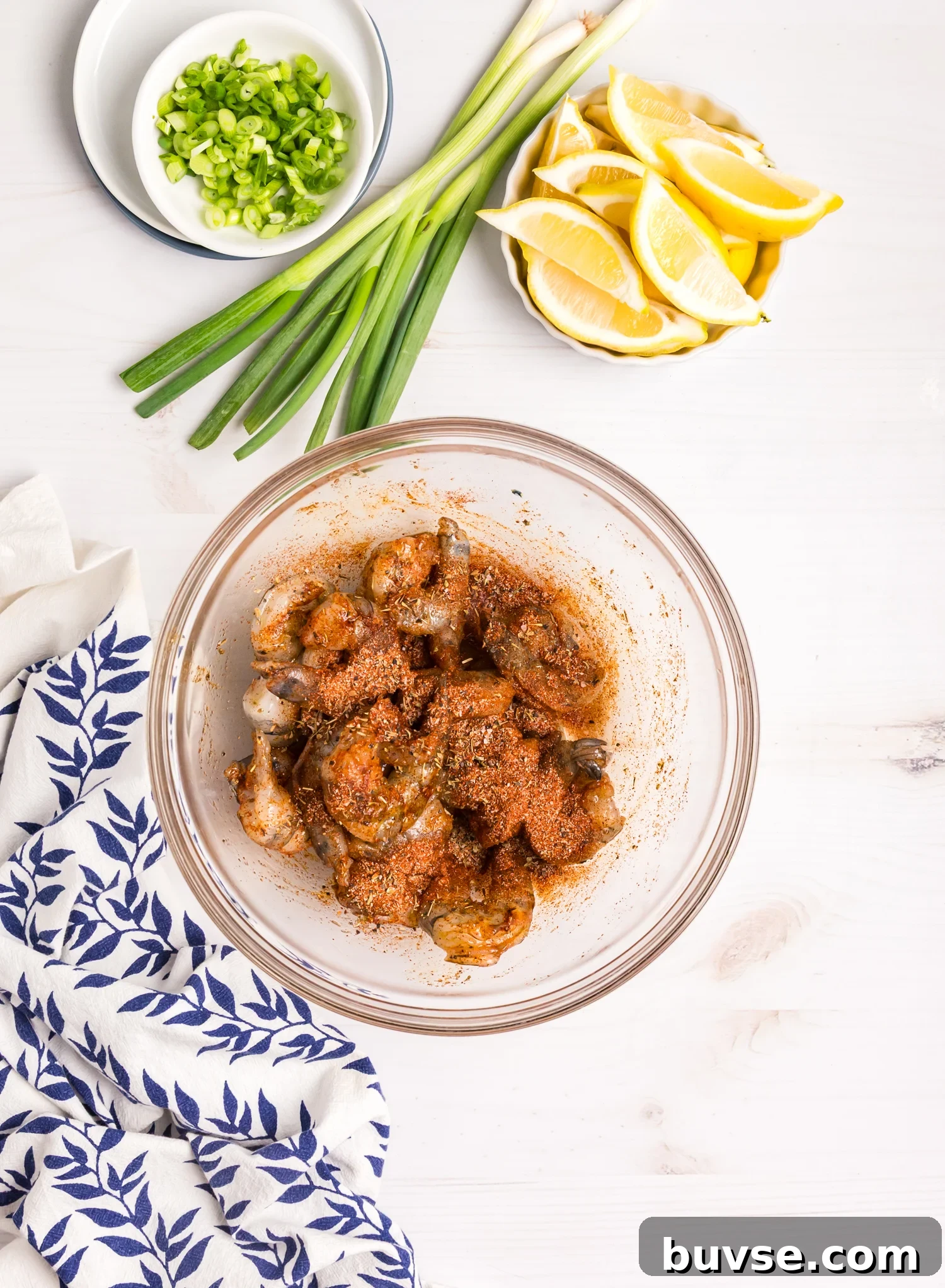 Seasoned raw shrimp in a bowl, coated evenly with spices, surrounded by fresh scallions and lemon slices.
