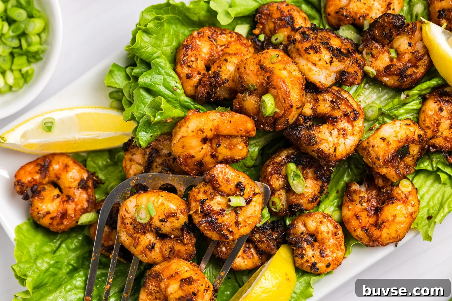 Close-up of grilled blackened shrimp on a bed of lettuce, garnished with sliced green onions and lemon wedges, with a fork lifting shrimp.