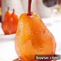 Port wine poached pear glistening in rich syrup on a white dessert plate, with more pears and red wine in the blurred background.