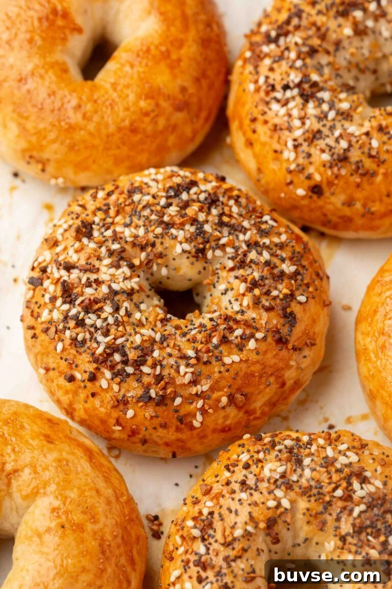 Golden gluten-free bagels, some topped with sesame seeds and seasoning, rest on a white surface. The bagels have a glossy finish and are freshly baked.