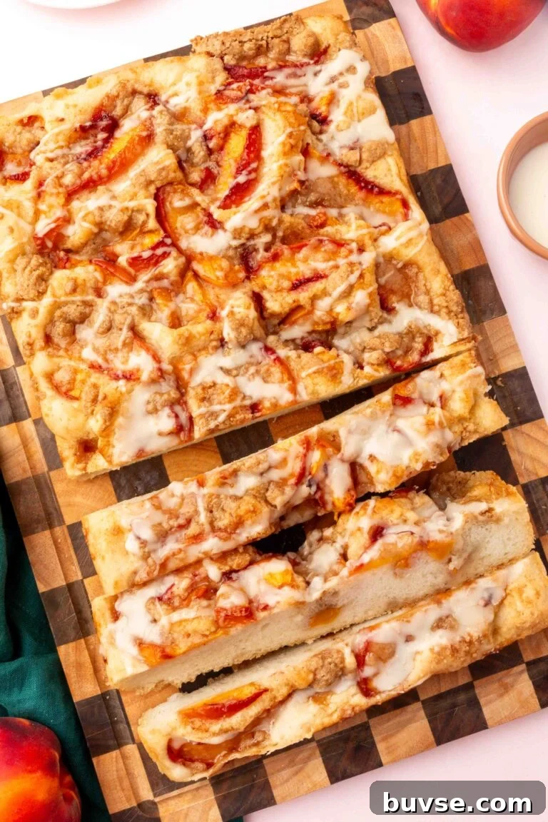 Rectangular Gluten-Free Peach Crumble Focaccia coffee cake with icing, sliced on a checkered wooden board. Cake has visible peach slices and streusel topping, surrounded by peaches and a small dish of icing.