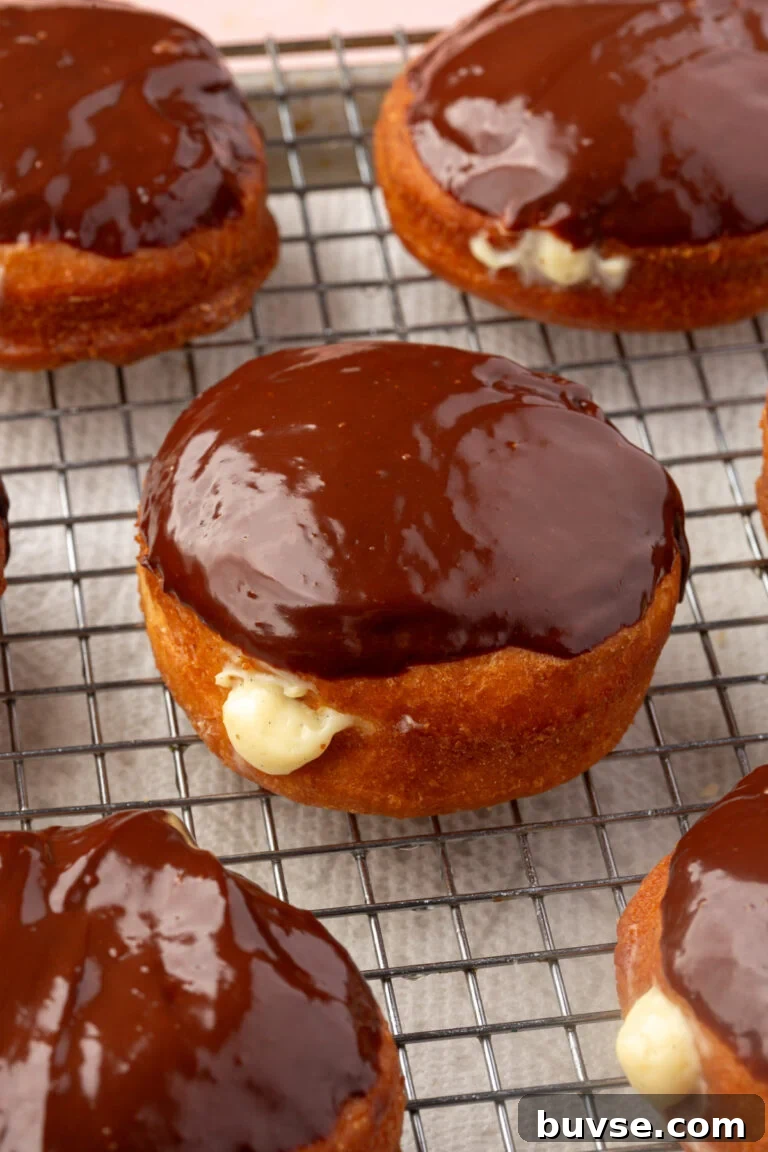 Boston cream donuts with chocolate glaze and creamy filling are cooling on a wire rack lined with parchment paper.