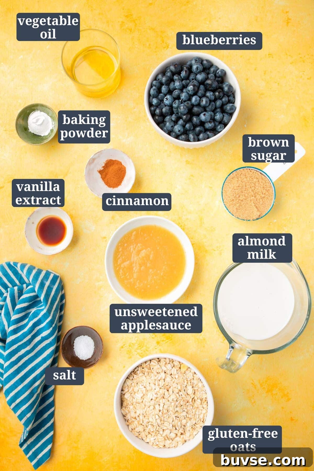 Blueberry Baked Oatmeal Recipe 3 Small bowls of ingredients on a yellow table to make blueberry baked oatmeal, including vegetable oil, baking powder, vanilla extract, brown sugar, unsweetened applesauce, almond milk, cinnamon, and gluten-free oats.