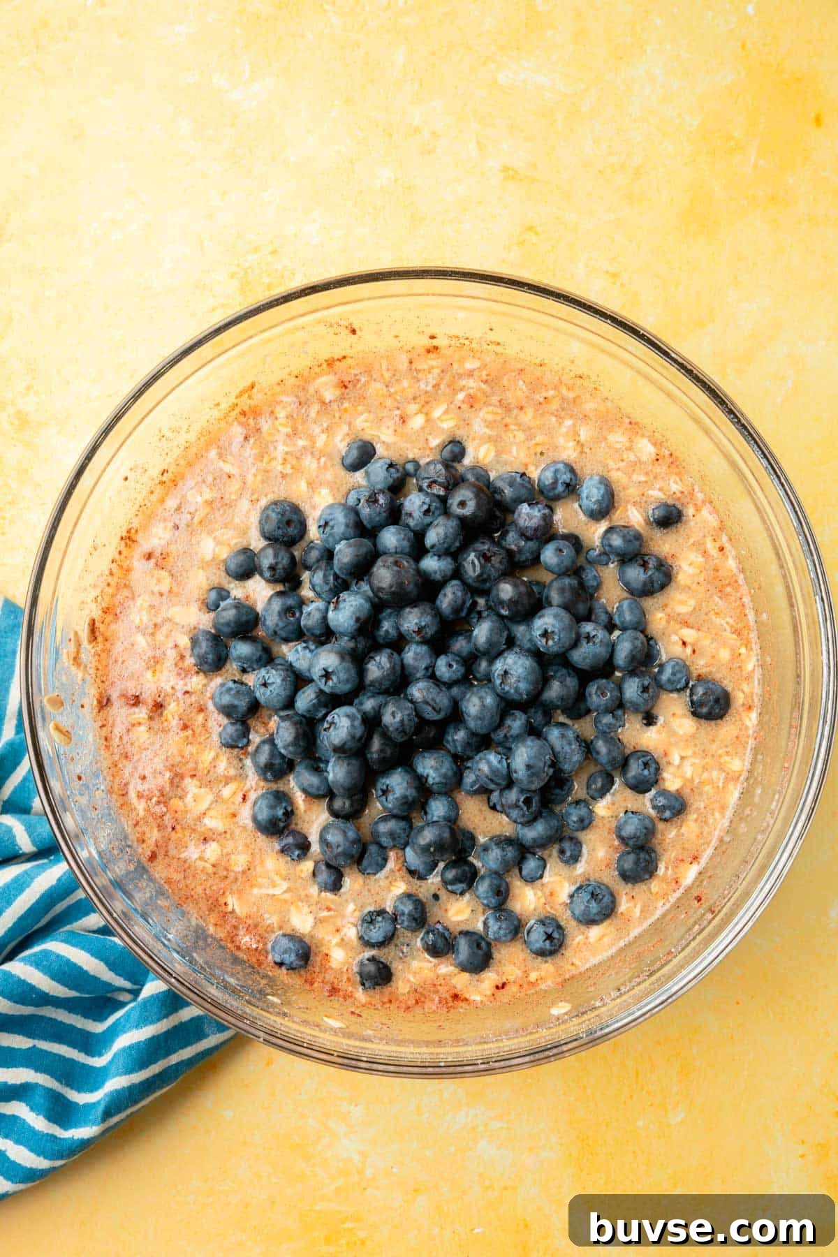 Blueberry Baked Oatmeal Recipe 7 A glass mixing bowl of a wet mixture of oats, almond milk, cinnamon, and applesauce topped with fresh blueberries that have not yet been mixed in.