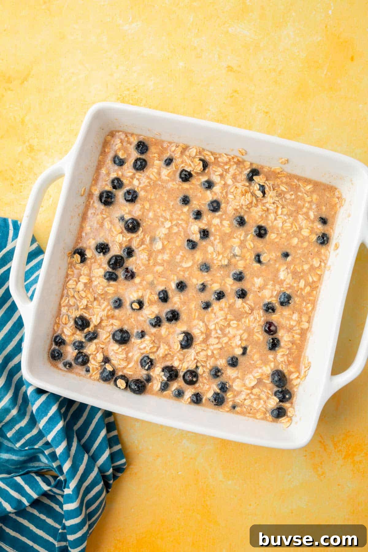 Blueberry Baked Oatmeal Recipe 8 A wet mixture of almond milk, spices, oats, and blueberries in a square baking dish on a yellow table.