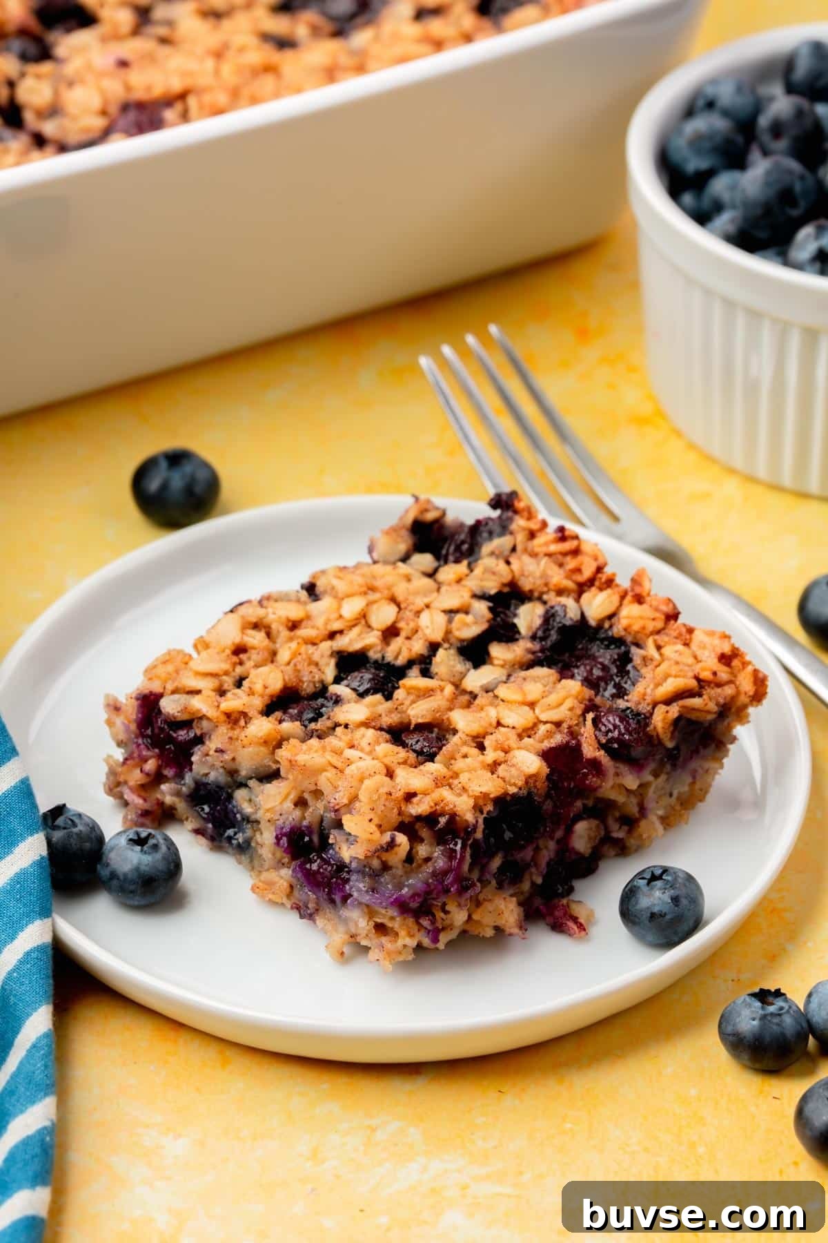 Blueberry Baked Oatmeal Recipe 10 A square of blueberry baked oatmeal on a small plate with a bowl of blueberries and a baking dish of baked oatmeal in the background.